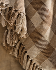 Checked natural throw with tassels draped over brown textiles.