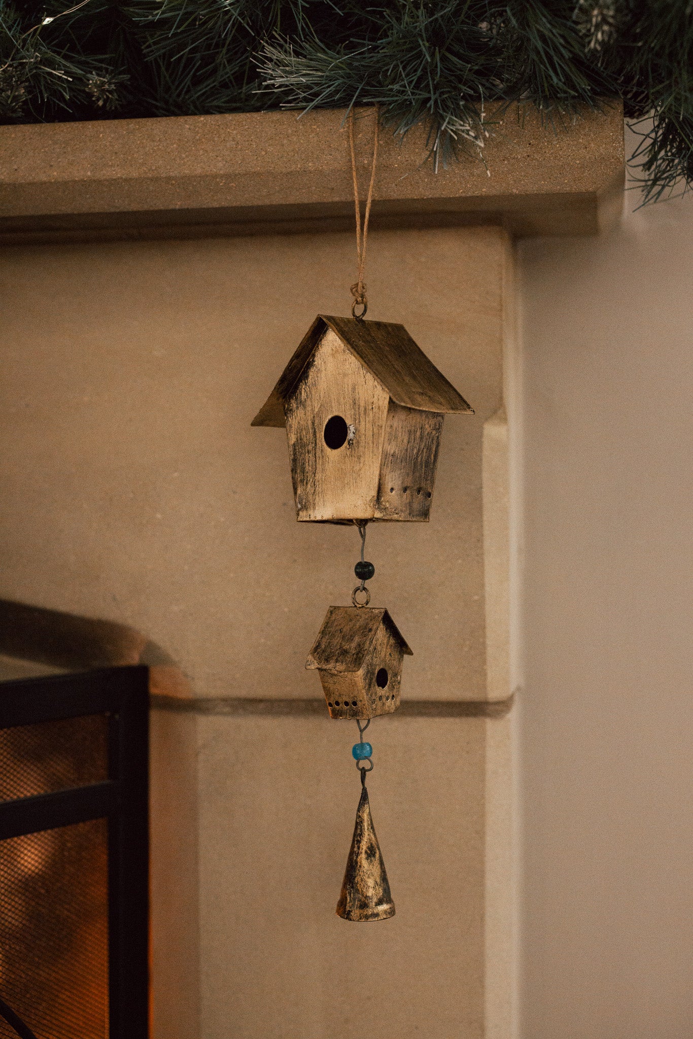 Antiqued brass bird house hanging decoration with two houses, a bell and green beads on stone fire place.