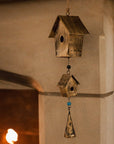 Antiqued brass bird house hanging decoration with two houses, a bell and green beads on stone fire place.