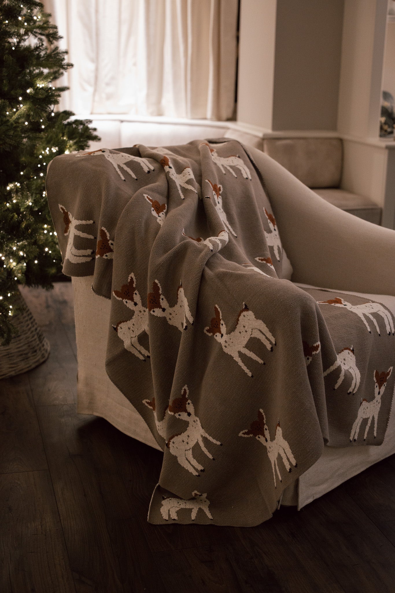 Neutral Christmas throw with Bambi reindeer print folded up on the arm of an armchair in front of Christmas Tree.