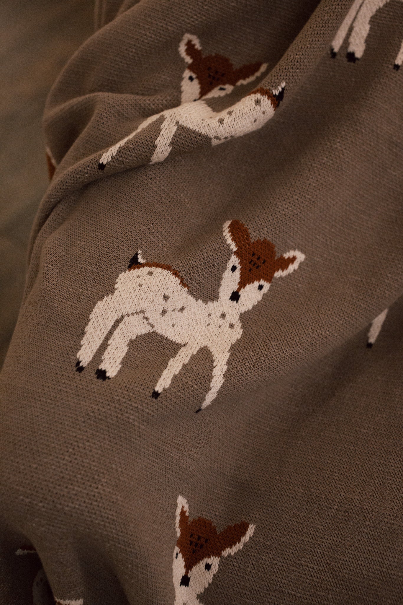 A close up of Neutral Christmas throw with Bambi reindeer print folded up on the arm of an armchair in front of fireplace.