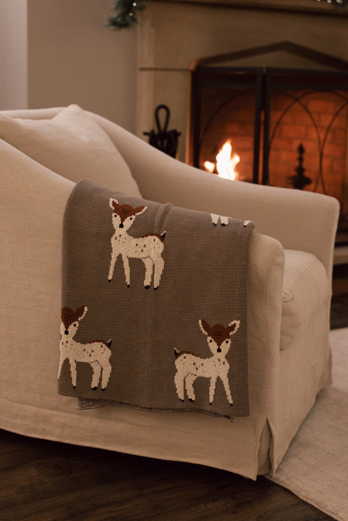 Neutral Christmas throw with Bambi reindeer print folded up on the arm of an armchair in front of fireplace.