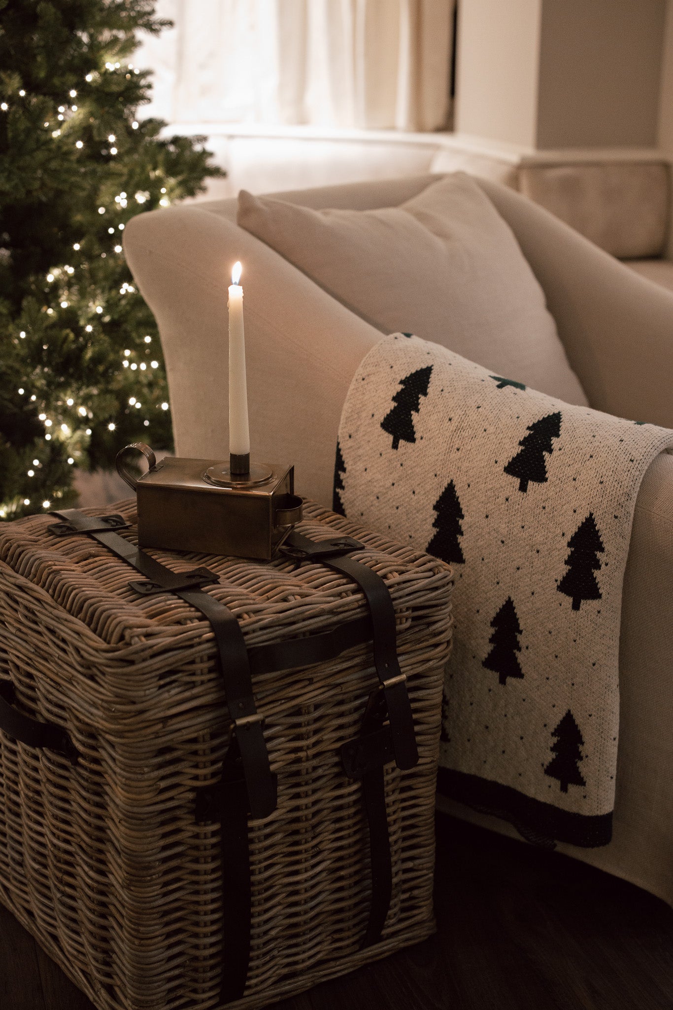 A rattan storage box side table with candle stick and christmas tree throw in front of lit up christmas Tree.