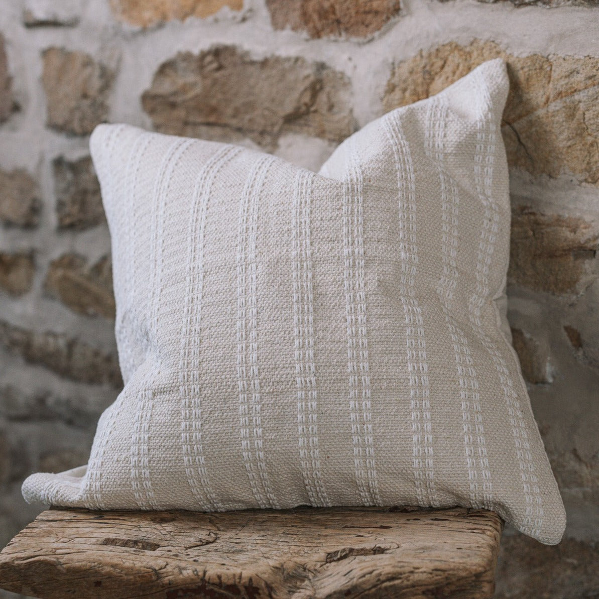 Cream and white stitch striped cushion on wooden stool against a stone wall.