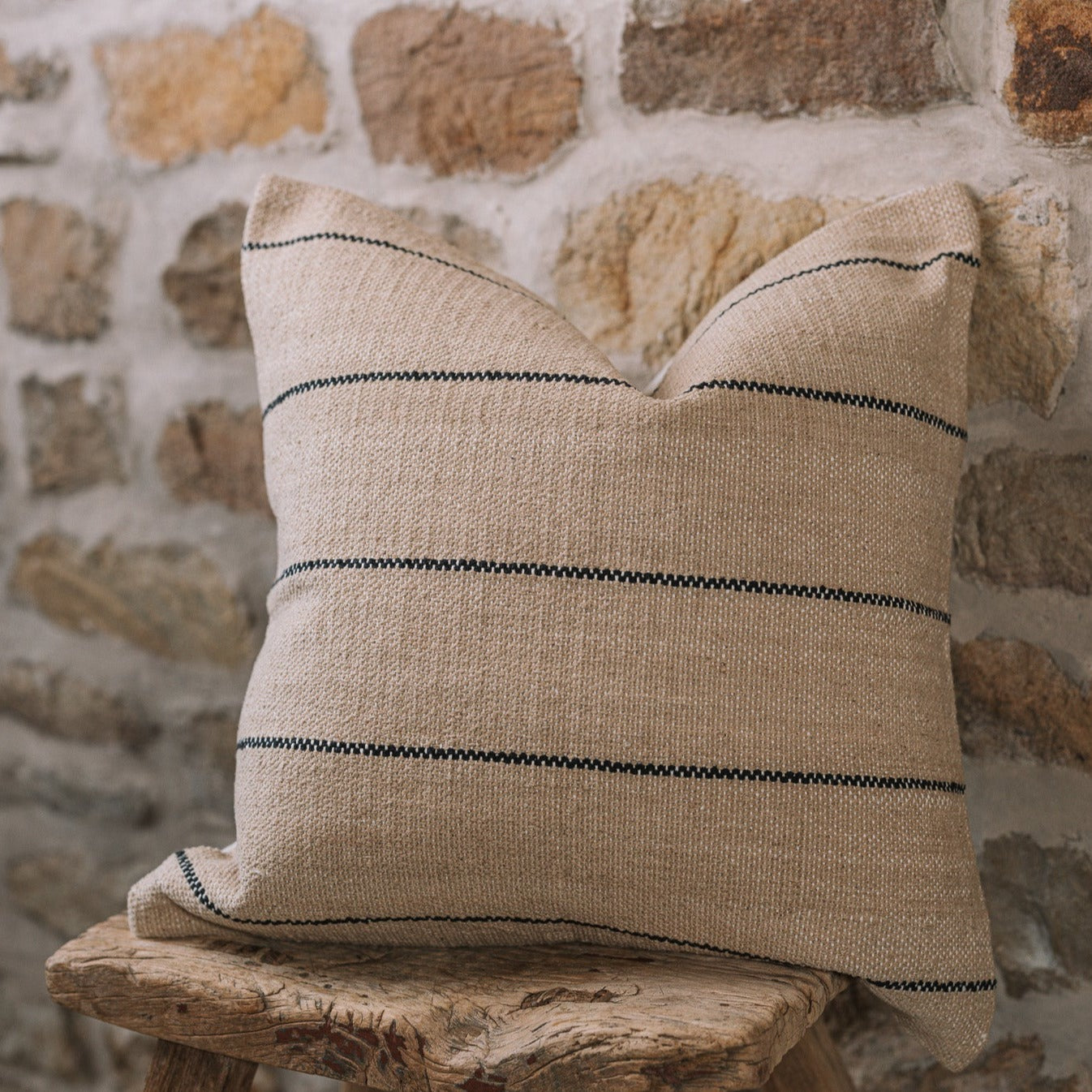 Natural cushion with black stripes on wooden stool against stone wall.