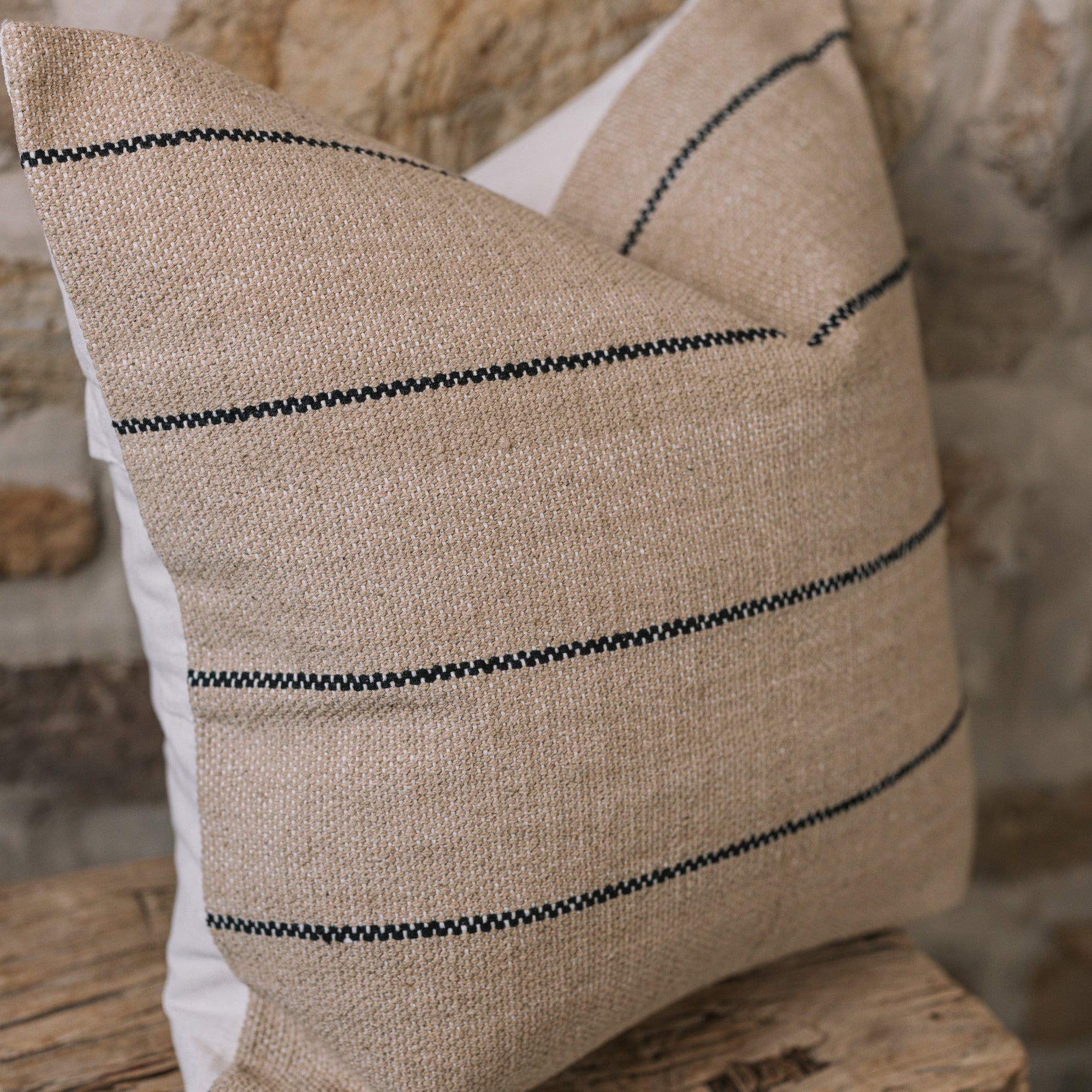 Natural cushion with black stripes on wooden stool against stone wall.