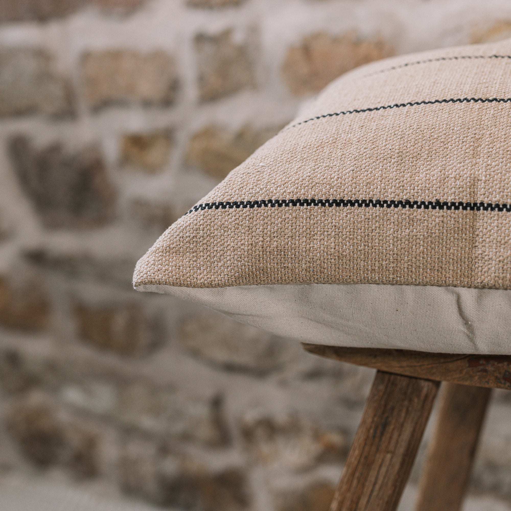 Natural cushion with black stripes on wooden stool against stone wall.