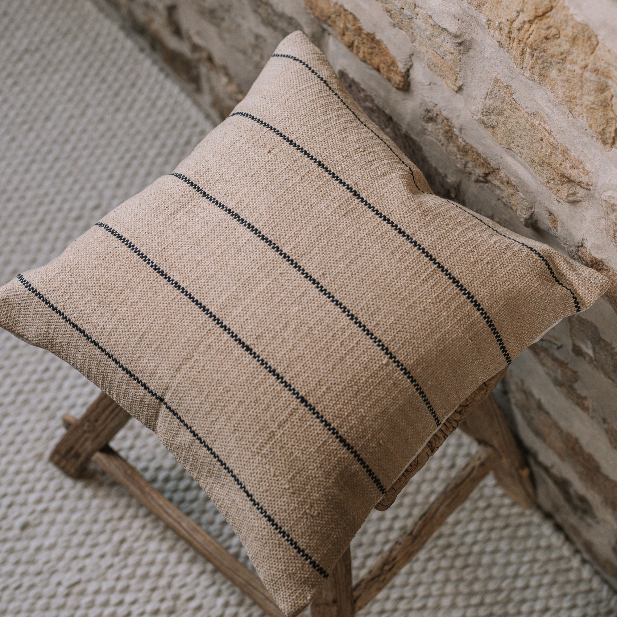 Natural cushion with black stripes on wooden stool against stone wall.