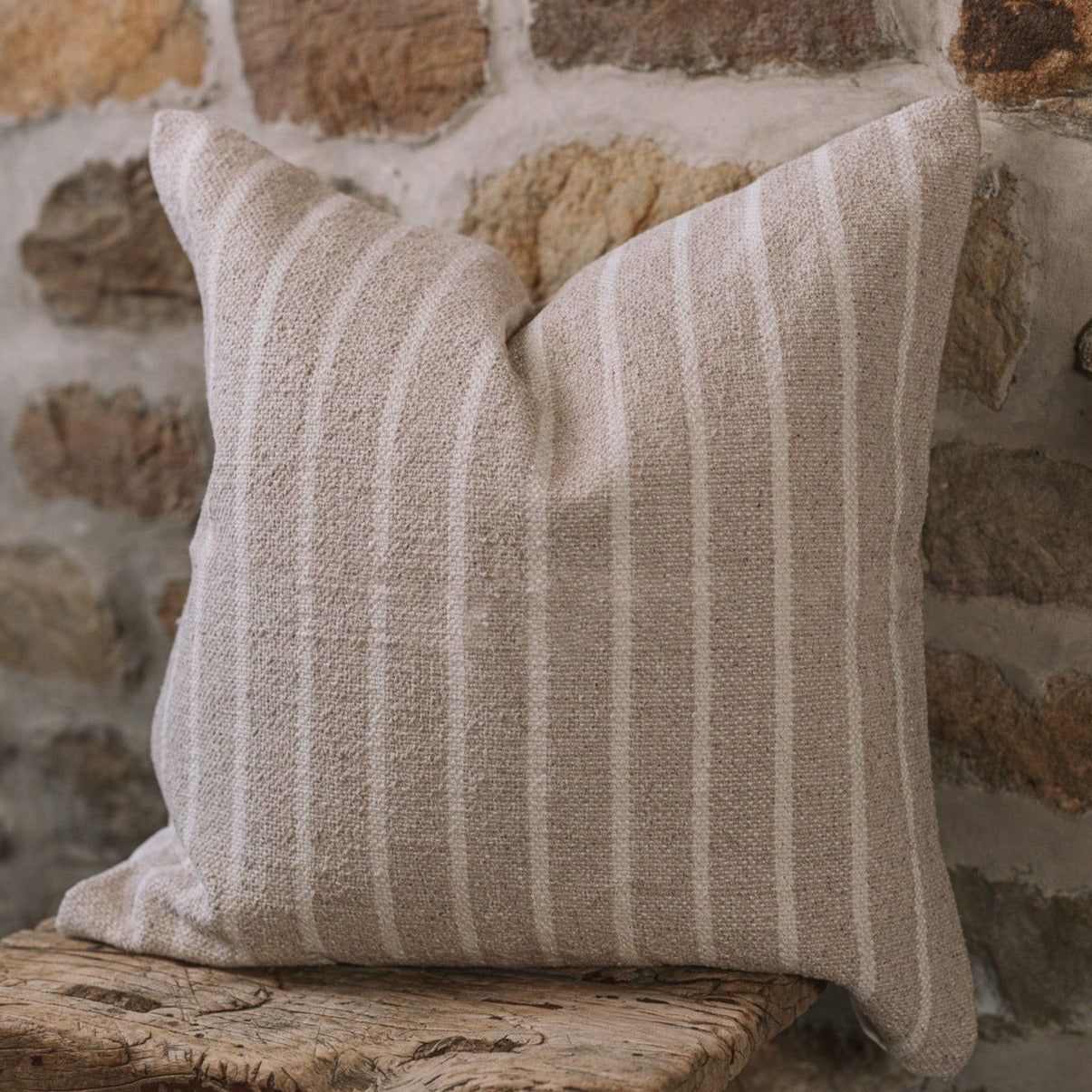 Light beige and cream stripe cushion on a wooden stool against a stone wall.