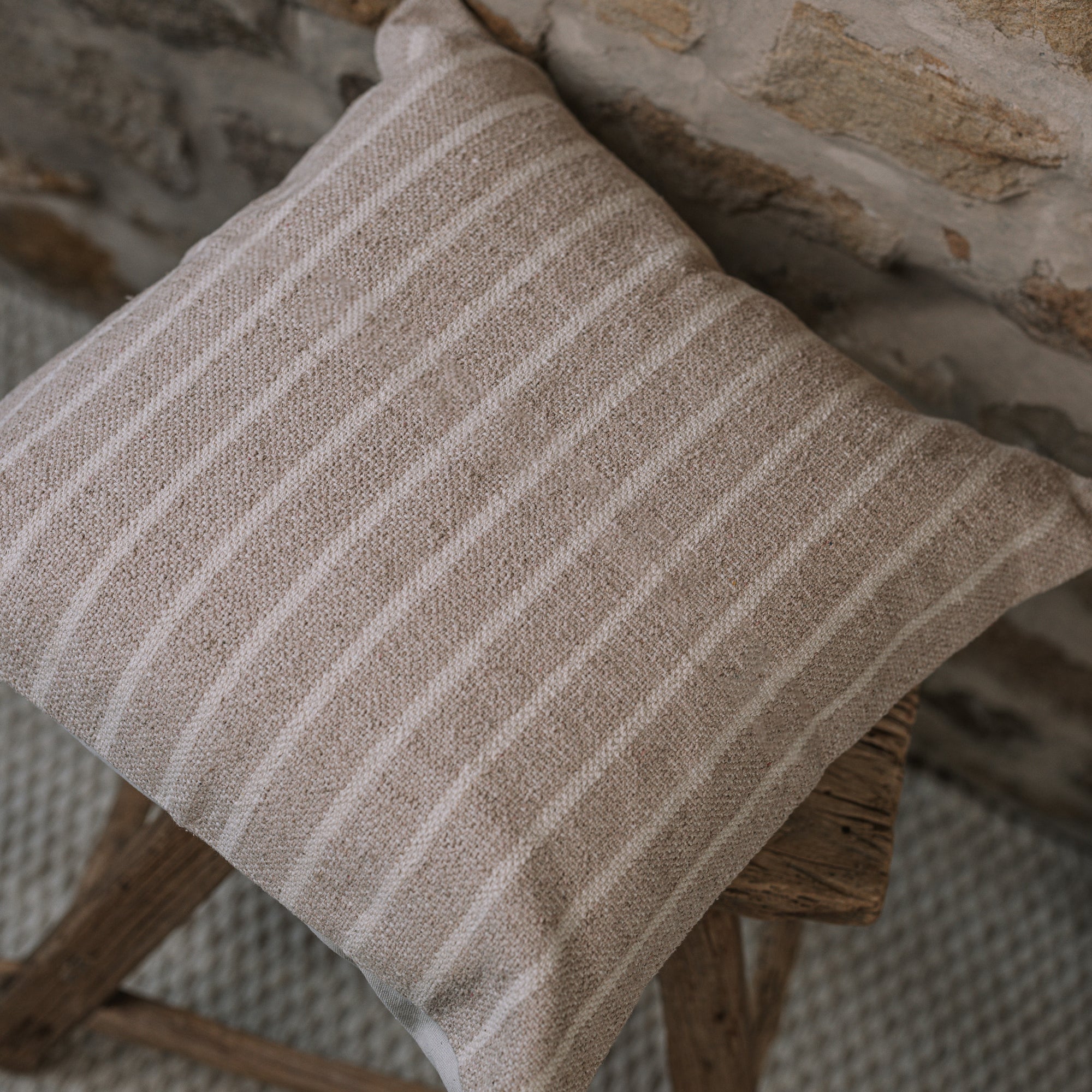 Light beige and cream stripe cushion on a wooden stool against a stone wall.