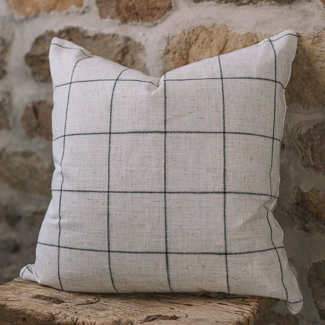 White and black grid cushion on wooden stool against a stone wall.