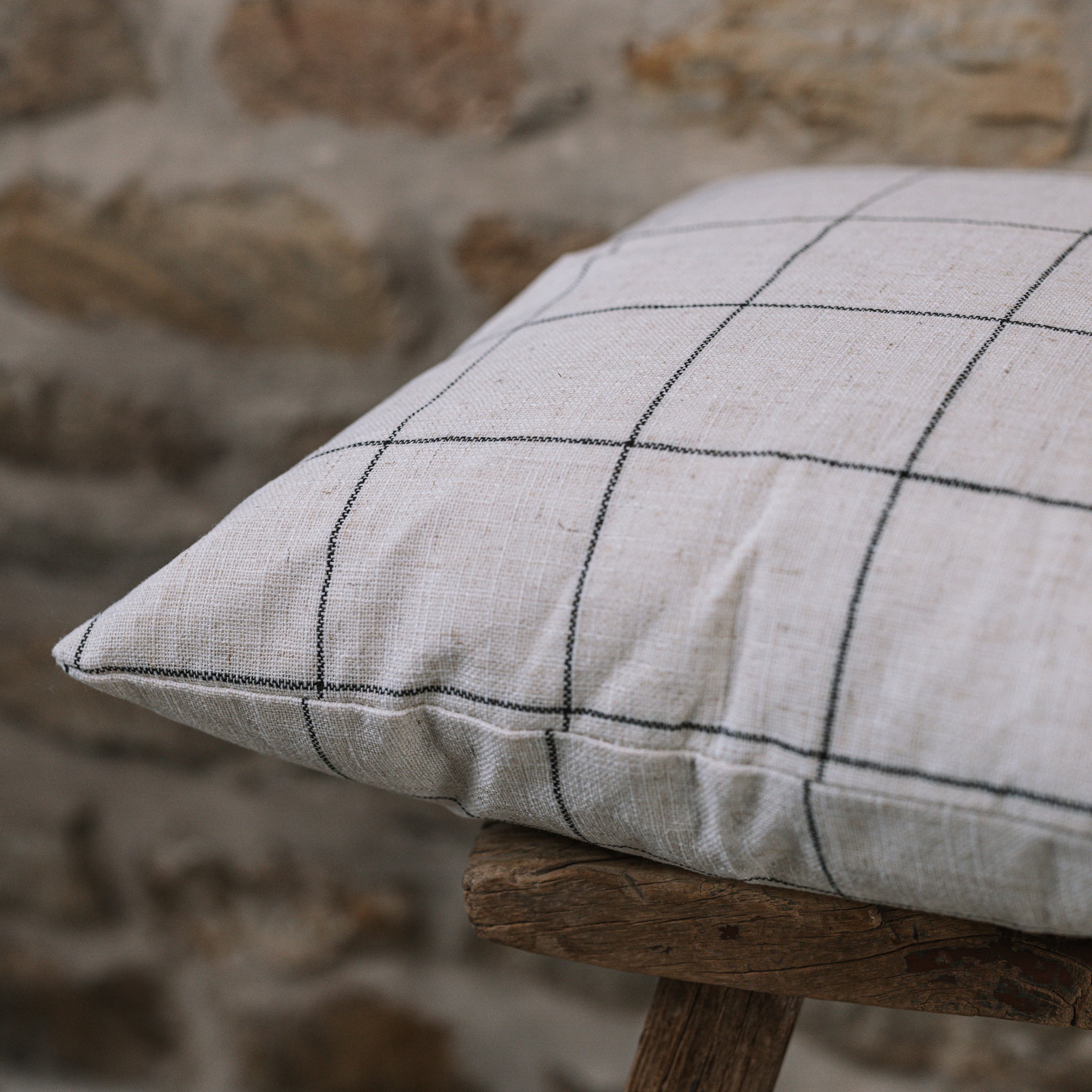 White and black grid cushion on wooden stool against a stone wall.