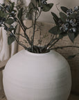 Ceramic round white textured vase with berry sprays and branches on a woven armchair.