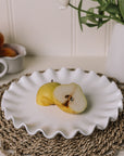 White ceramic cake plate with ruffled edges with a sliced pear on a rattan placemat.