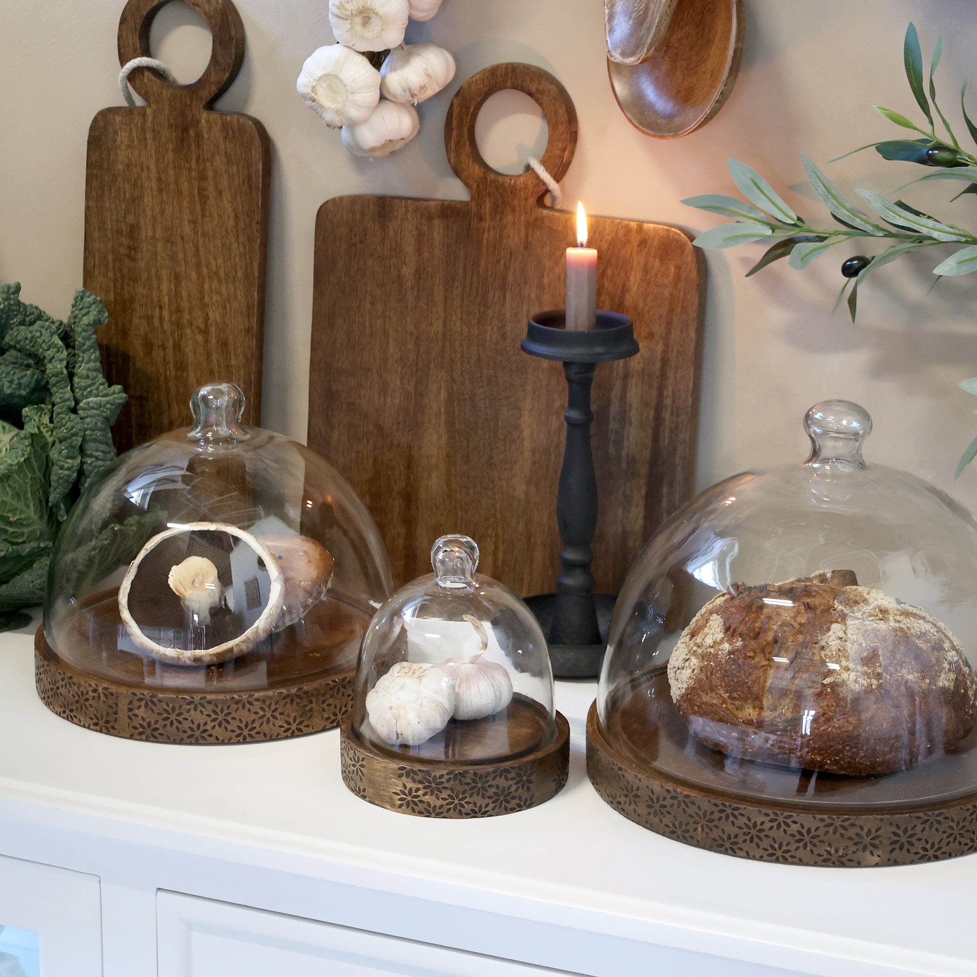 3 wooden and glass domes with food inside, on a kitchen counter top with a candle stick and chopping boards.