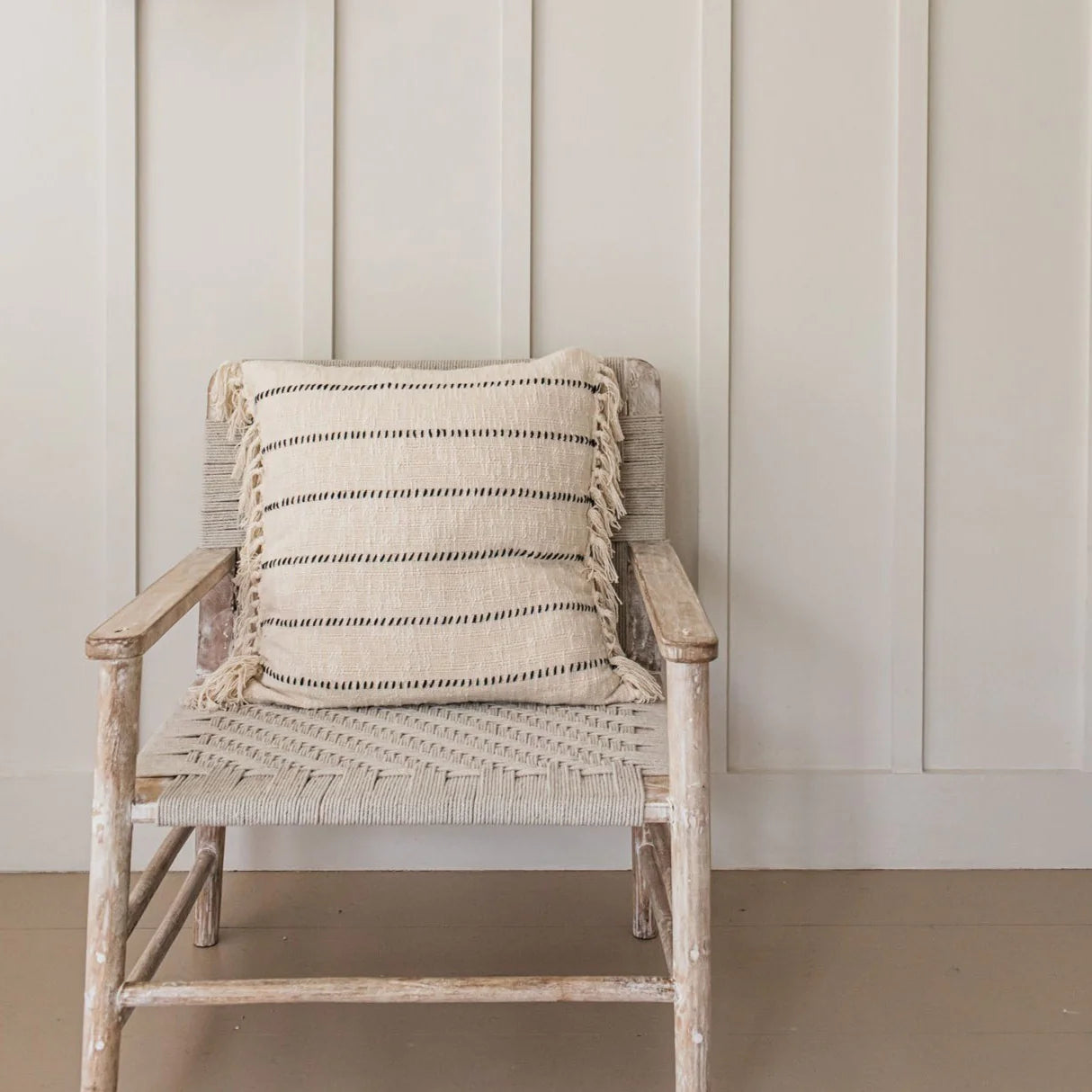 Striped neutral cushion sits on a woven armchair against a cream panelled wall.