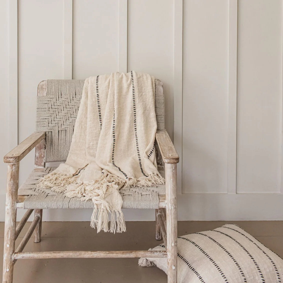 A striped neutral throw blanket is draped over a woven armchair with a matching cushion on the floor.