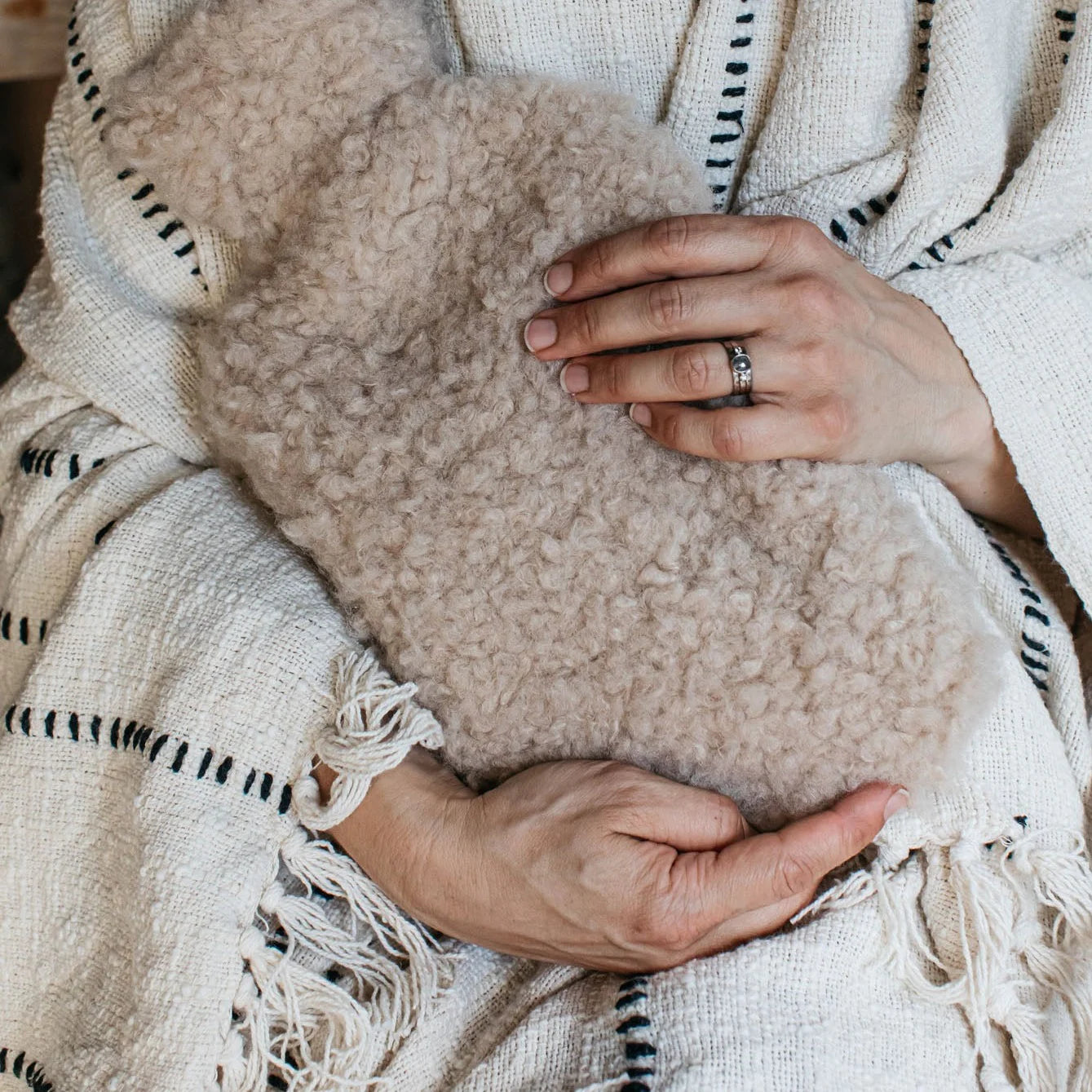 A woman holds a sheepskin hot water bottle, she has a striped neutral throw blanket wrapped around her. 