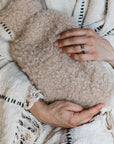 A woman holds a sheepskin hot water bottle, she has a striped neutral throw blanket wrapped around her. 