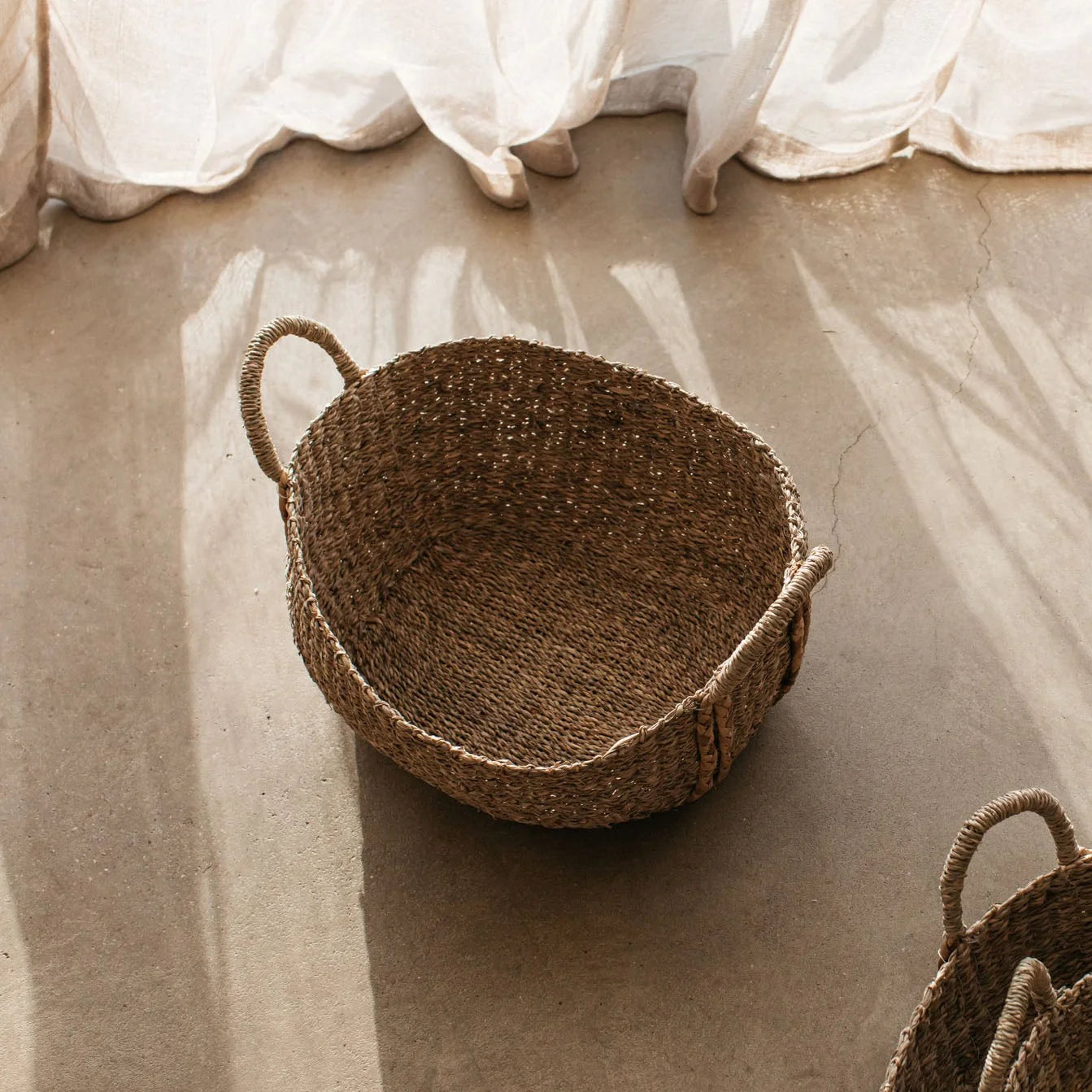 The Audrey oval seagrass basket on a cement floor with linen curtain in background.
