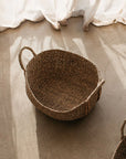The Audrey oval seagrass basket on a cement floor with linen curtain in background.