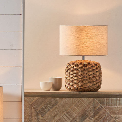 Small woven table lamp with a light lamp shade switched on, on a wooden console with two cups.