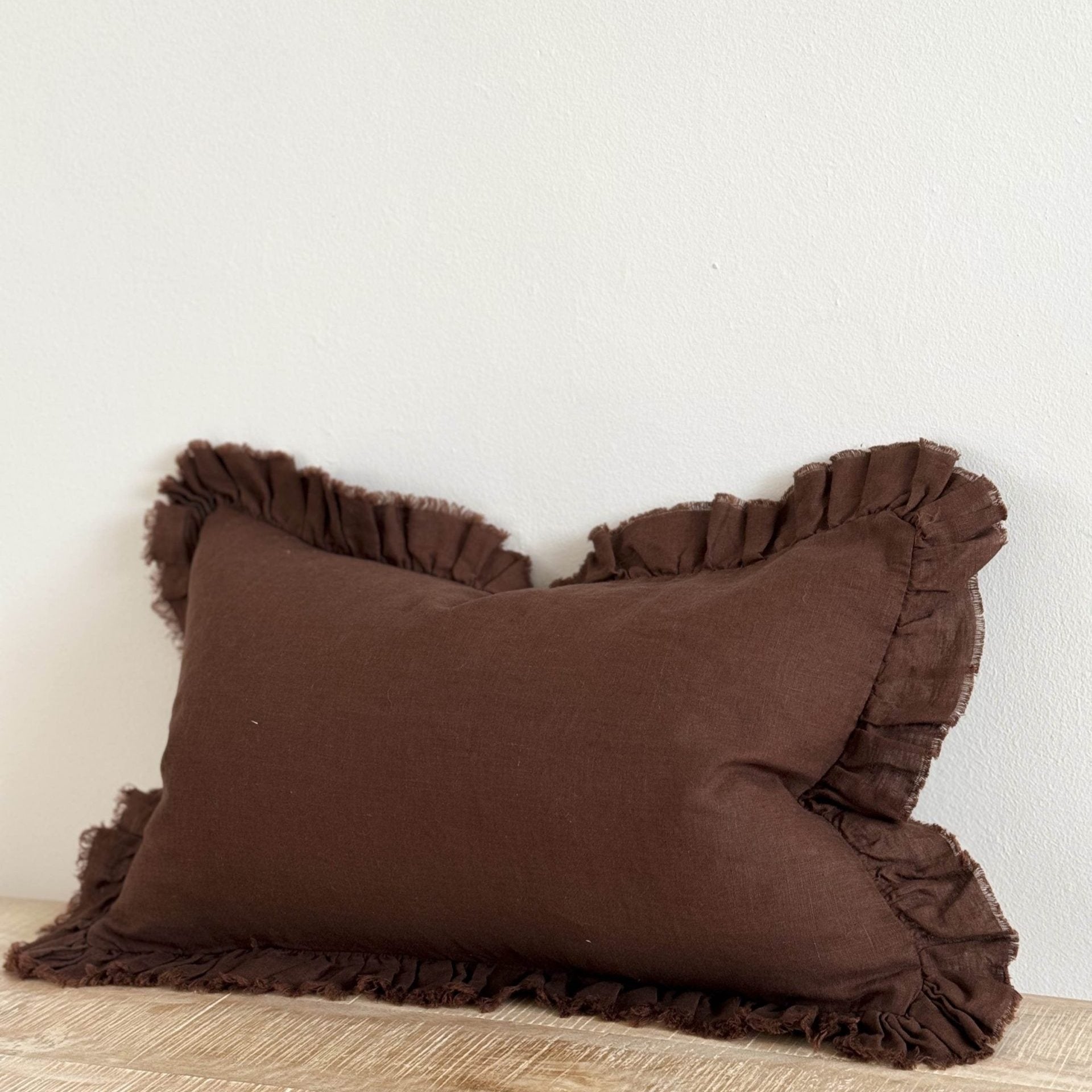 Chocolate brown rectangle linen cushion with ruffle edge on wooden surface.