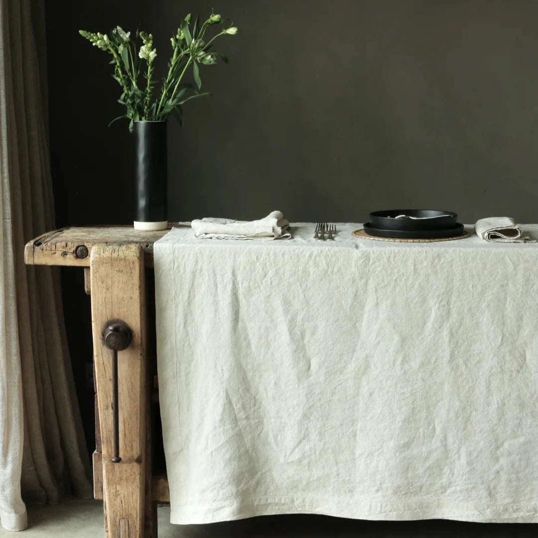 Natural linen table cloth draped over table with dinner set, cutlery and vase.