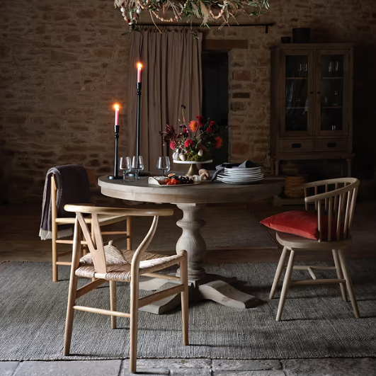 Wooden round dining table in large stone room, with candelabras, cushions and tableware.