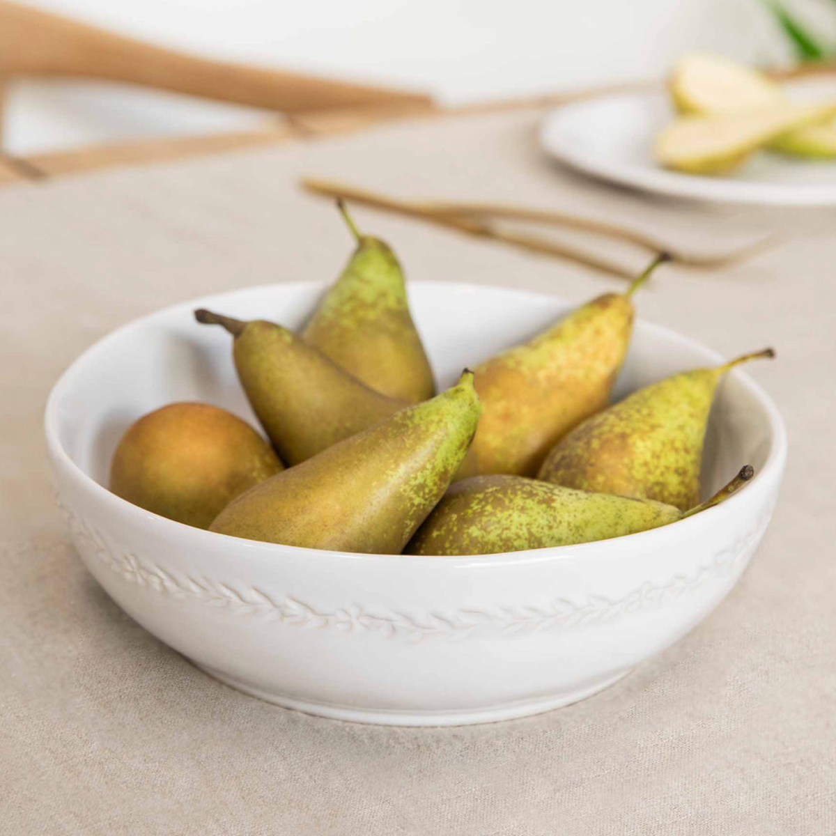 White ceramic serving bowl with leaf detailing, filled with pears on dining table.