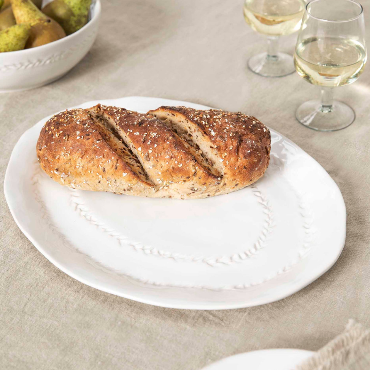 White ceramic serving platter with leaf detailing, loaf of bread on top and glasses of wine aside.