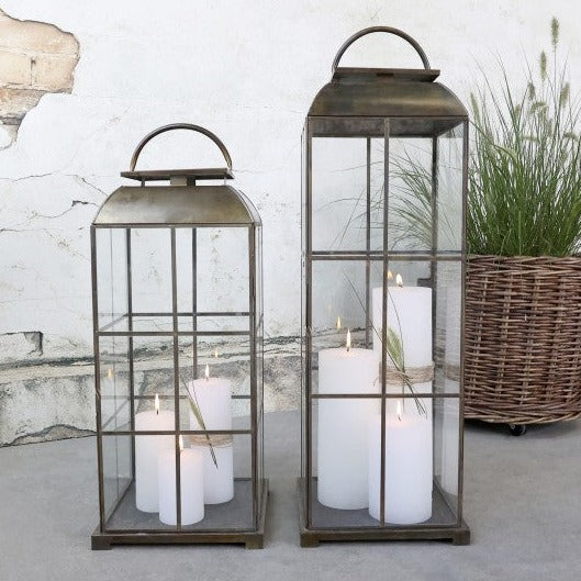 Set of to brass lanterns filled with lit white candles in front of a plant.