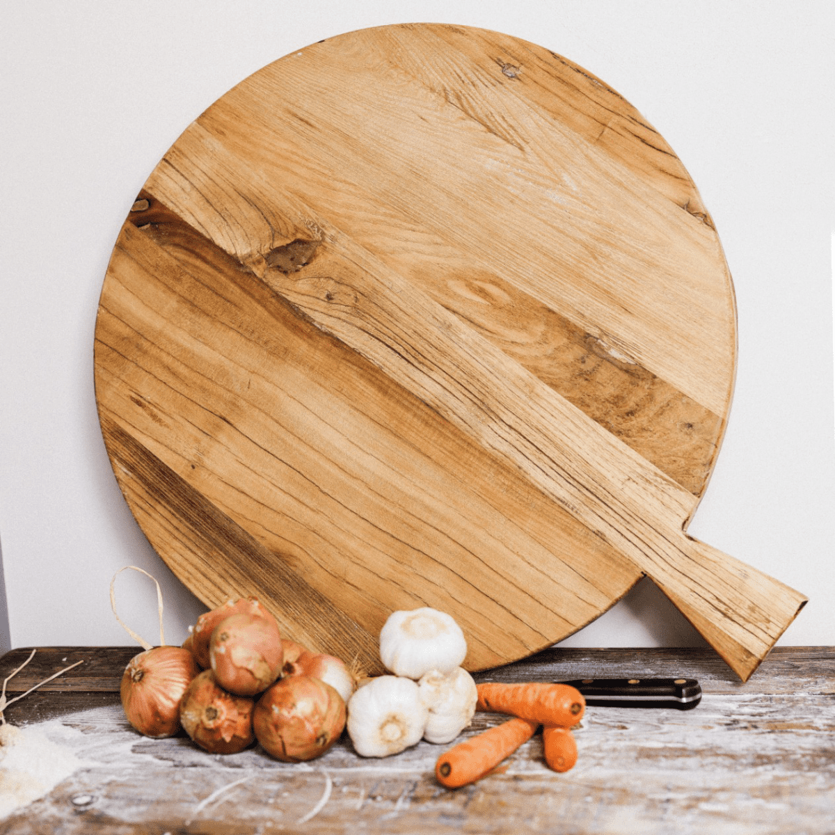 Circular wooden chopping board with handle against a wall with vegetables and a knife in front.