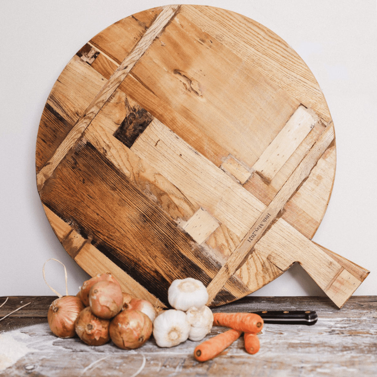 Back of circular wooden chopping board with handle against a wall with vegetables and a knife in front.
