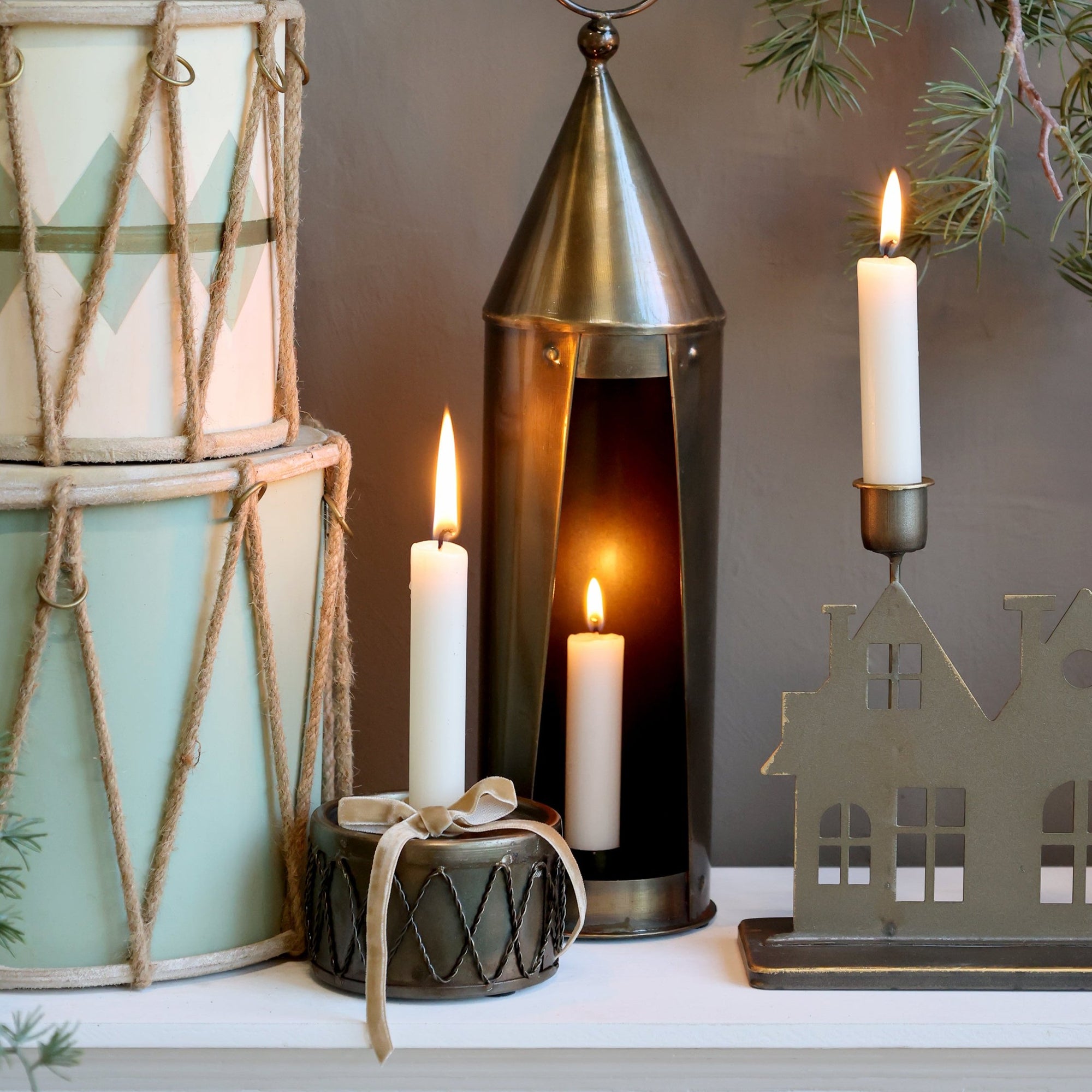 Drum shaped candlestick holder with lit candle inside, a bow and other surrounding Christmas decorations.