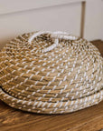Woven seagrass cake dome with white loops on top, on wooden console.