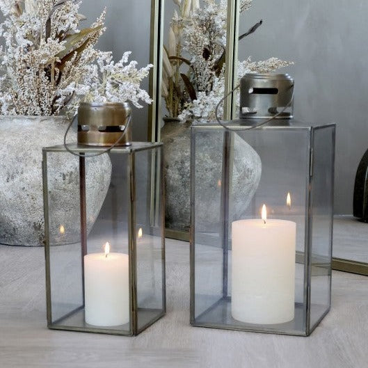 Two glass and brass candle lanterns with lid candles inside and plant behind.