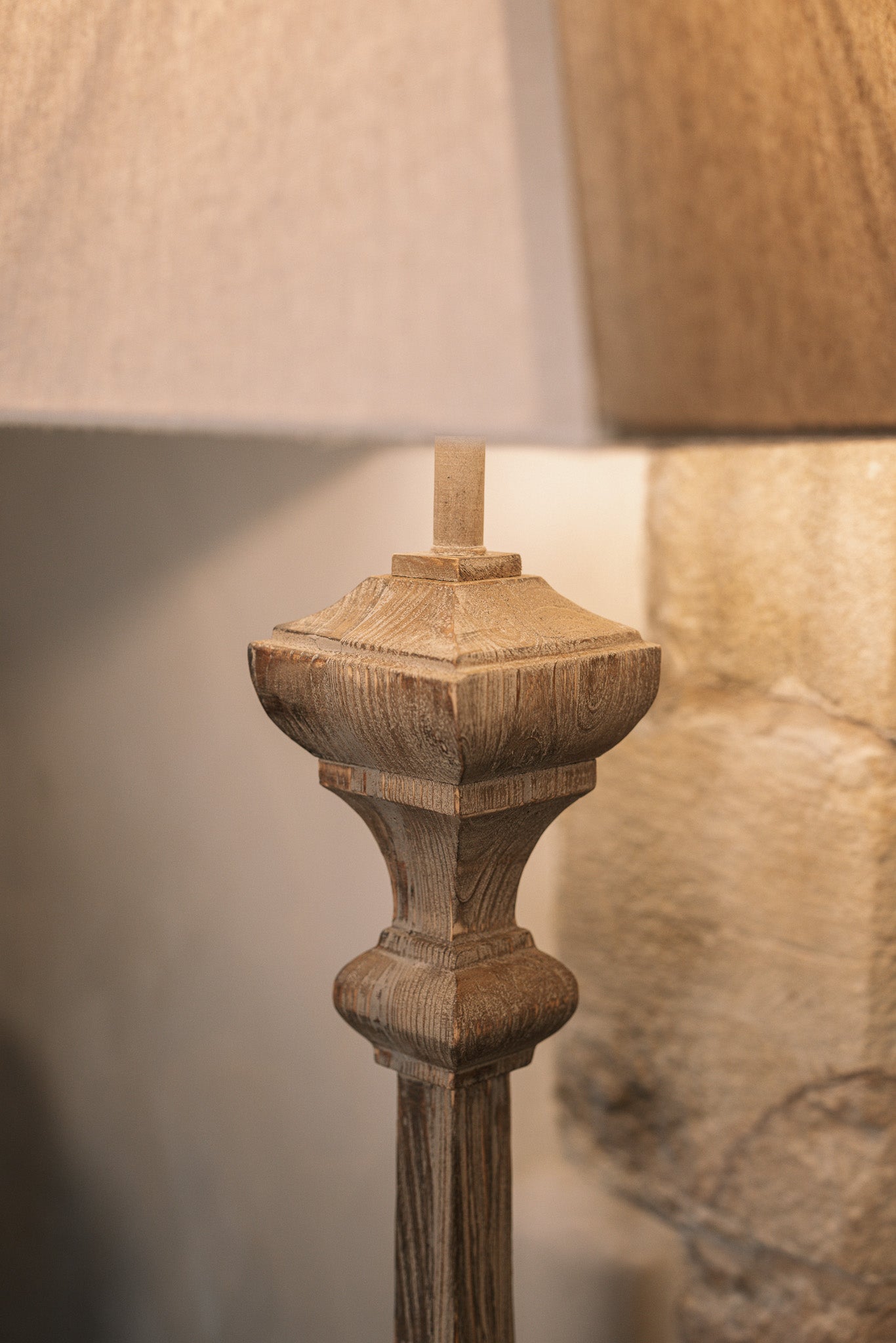 Carved wooden floor lamp with white linen shade in room with stone wall.