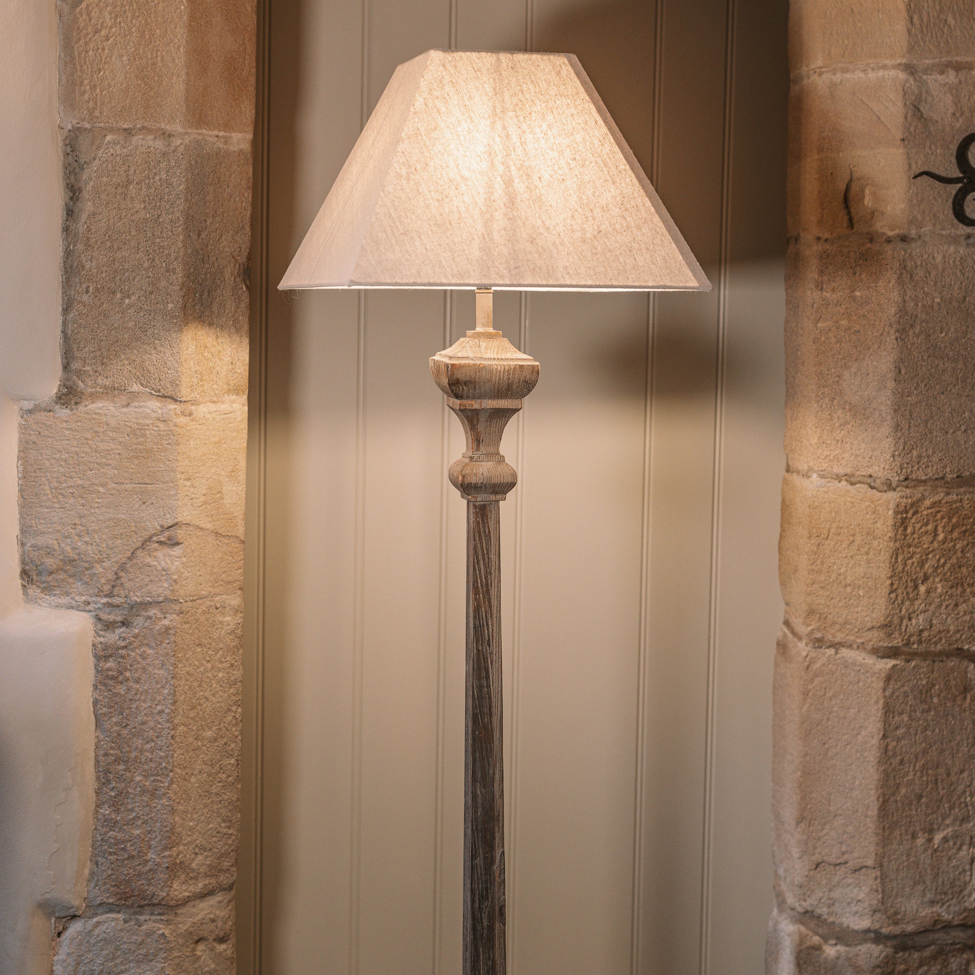 A carved wooden floor lamp with square linen shade in front of stone arch.