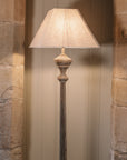 A carved wooden floor lamp with square linen shade in front of stone arch.