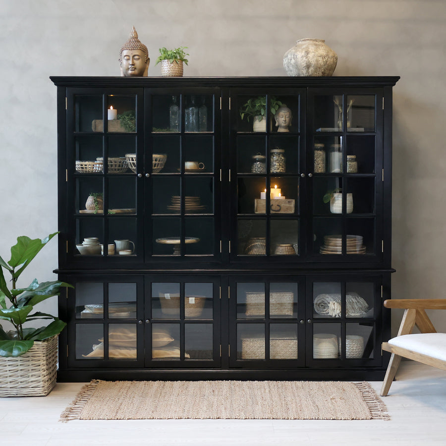 Large black armoire filled with home decor in neutral room with rug, plant and chair.