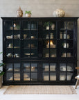 Large black armoire filled with home decor in neutral room with rug, plant and chair.