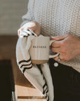 White and neutral brown stoneware mug with 'blessed' written in black text, being dried with tea towel.