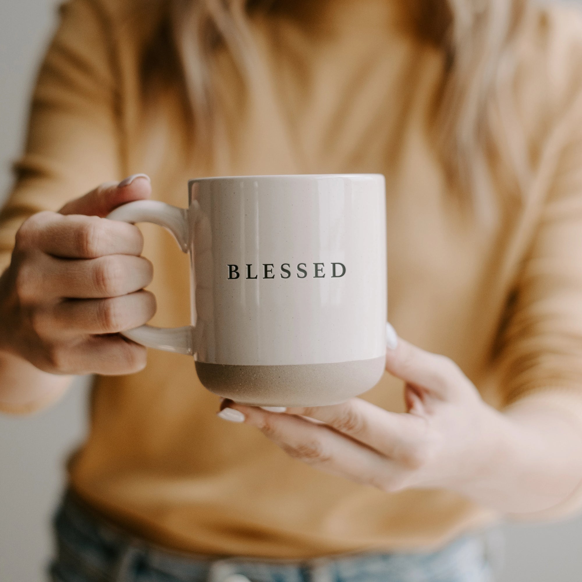 White and neutral brown stoneware mug with 'blessed' written in black text, held up with two hands.