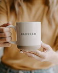 White and neutral brown stoneware mug with 'blessed' written in black text, held up with two hands.