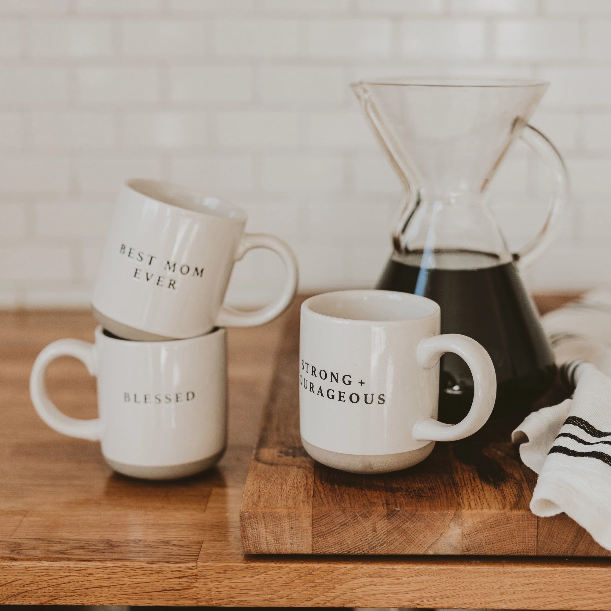 Selection of white and neutral brown stoneware mugs with slogans in black text, in front of cafetiere.