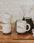 Selection of white and neutral brown stoneware mugs with slogans in black text, in front of cafetiere.