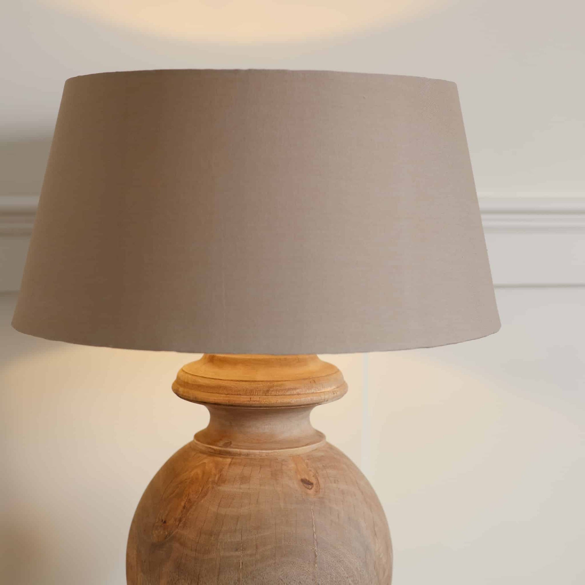 Beige linen lamp shade on round wood lamp, switched on in front of white background.