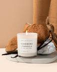 Cashmere and vanilla soy candle in white jar, lit in front of some coconuts and vanilla beans. 