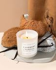 Cashmere and vanilla soy candle in white jar from above, lit in front of some coconuts and vanilla beans. 